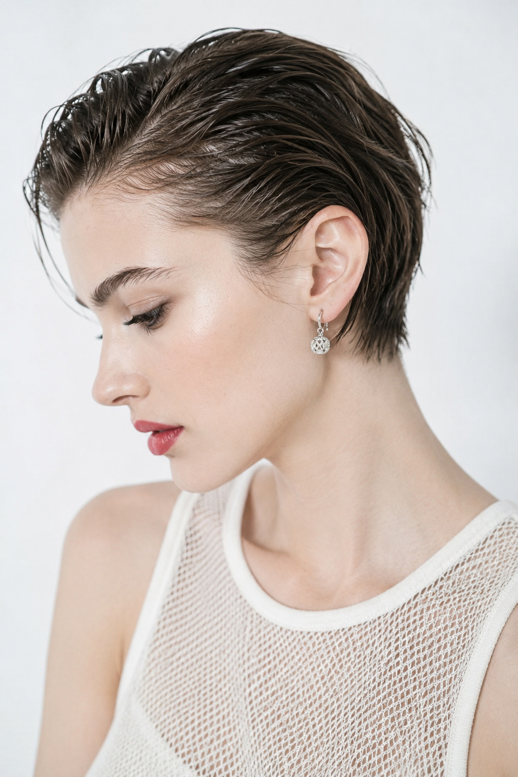 Upper body close-up of a female model with a textured, slightly messy wet-look pixie haircut in chocolate brown. Wearing delicate silver drop earrings and a sheer white summer mesh top. Against a plain off-white background. The model has an effortless, relaxed expression, looking downward slightly with a high-angle shot.