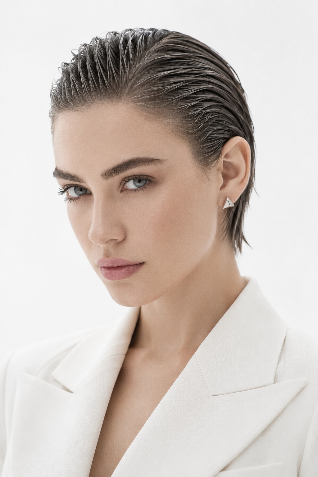 Upper body close-up of a model with a sleek, flat-combed wet look pixie haircut in charcoal grey. Decorated with elegant geometric silver studs and a structural, forward-fashion summer blazer. Set against a pure white minimalist background. The model has a neutral, high-fashion expression, captured in a direct close-up angle.