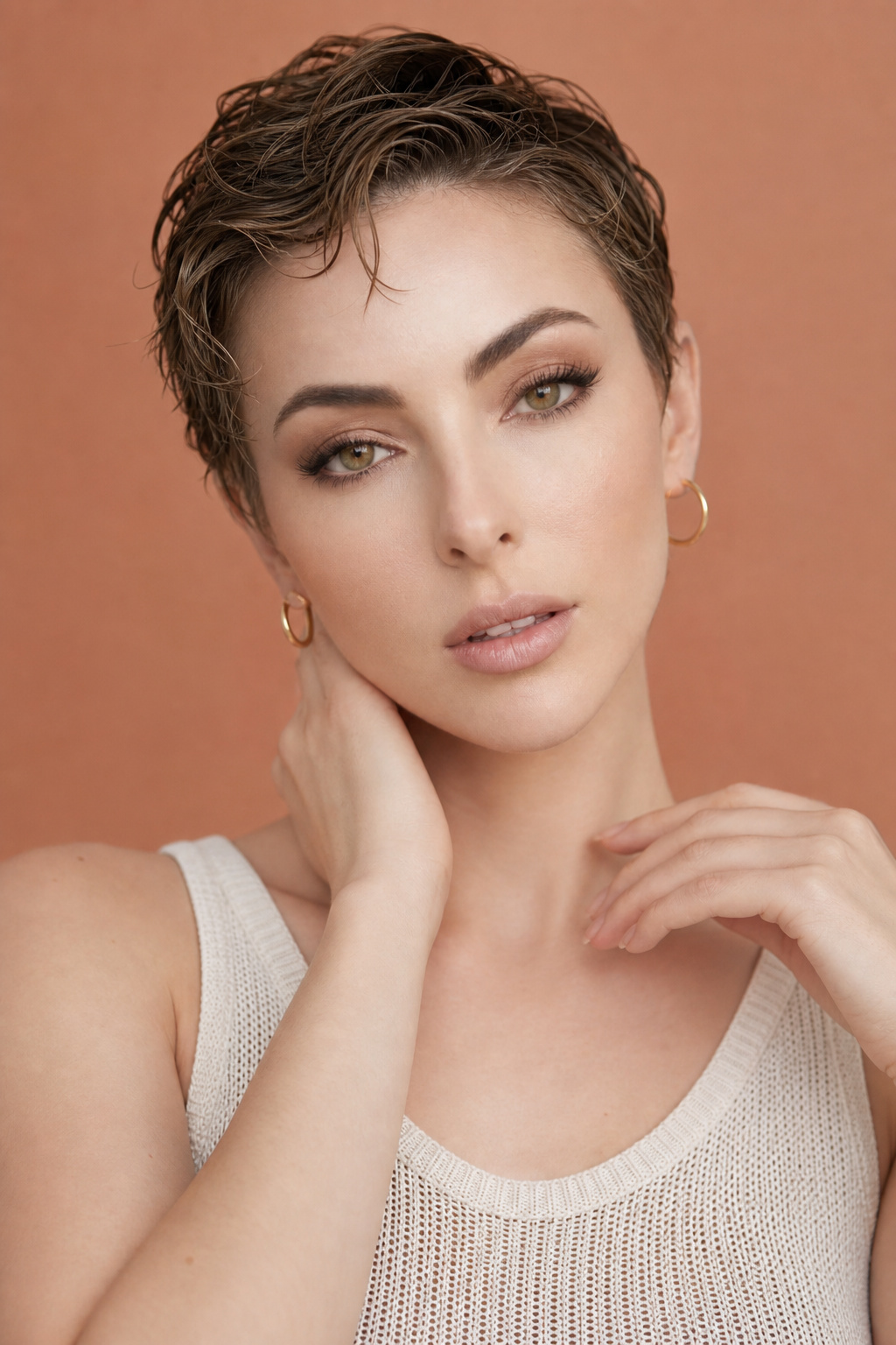 An upper body close-up of a female model with natural wavy wet look pixie hair in rich auburn. She wears delicate thin gold hoops and a summer knit tank top. Set against a minimalist warm terracotta background. The model has a soft, inviting expression, captured from a slightly elevated angle.