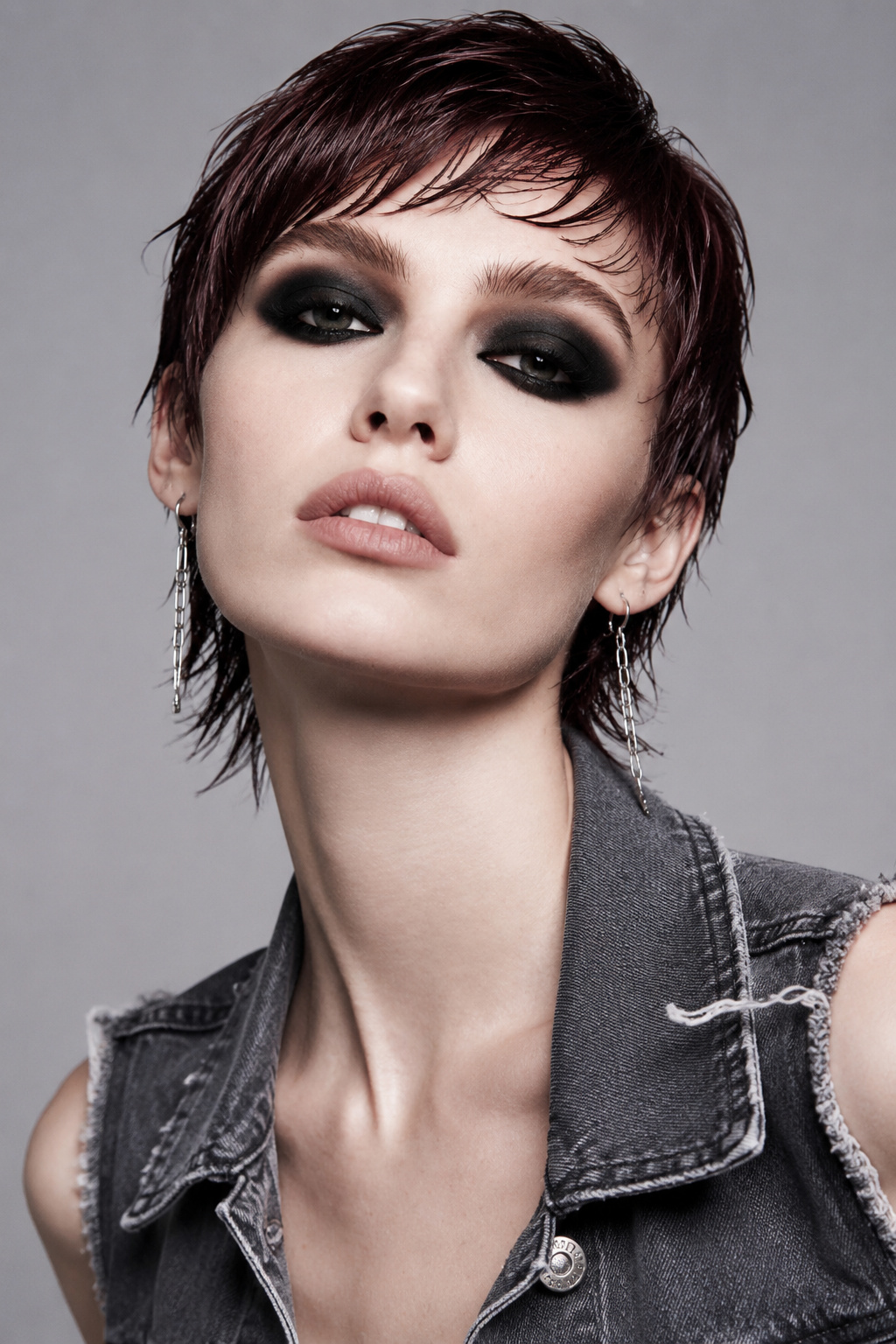 An upper body close-up of a model sporting a shaggy, mullet-inspired wet pixie cut in deep plum. Wearing delicate chain-link earrings and a sleeveless distressed denim vest. Minimalist charcoal gray background. The model shows a rebellious, confident expression, looking directly at the camera with a low-angle shot.
