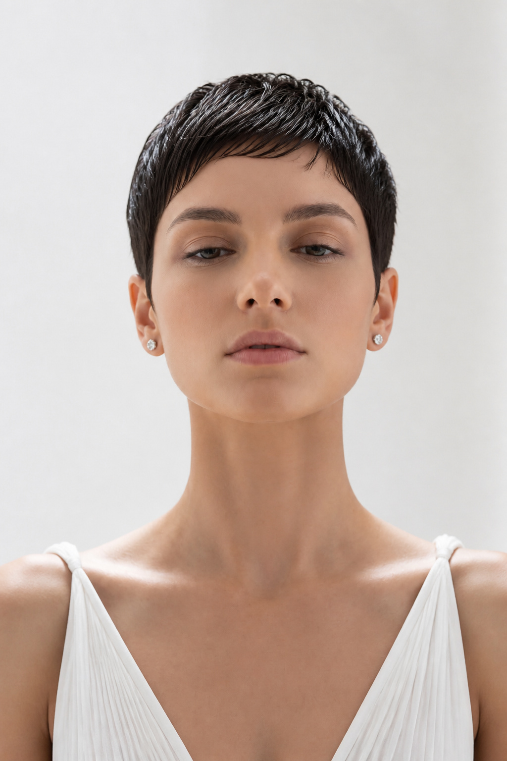 Luxuriöses Beauty-Porträt einer Frau mit einer schwarzen edgy Pixie Cut Frisur mit glänzendem Wet-Look-Finish und glatten Schichten. Sie trägt Diamant-Steckohrringe und ein modisches weißes Sommerkleid. Minimalistischer sauberer Hintergrund, eleganter Ausdruck, direkter Blickkontakt, High-End-Editorial-Haarstyling-Fotografie.